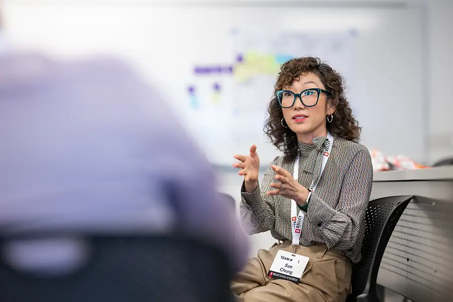 A Kelley School of Business online programs MBA student is in discussion with another student on a teamwork project. There is a whiteboard with writing on it in the background.